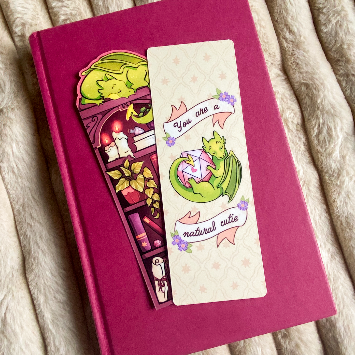 Picture of a fantasy dragon bookmark in a book. The dragon sleeps on top of the bookshelf that is filled with books and trinkets.