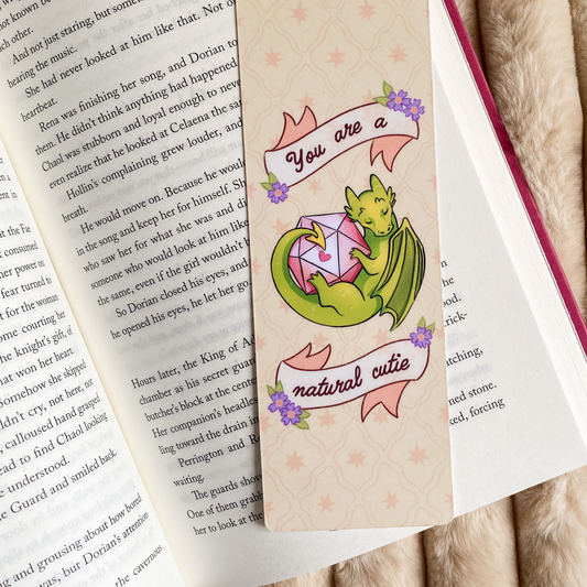 A product photo of a bookmark with an illustration of a green dragon holding a D20 dice. There is text saying ''you are a natural cutie''. The bookmark is laying in a book.