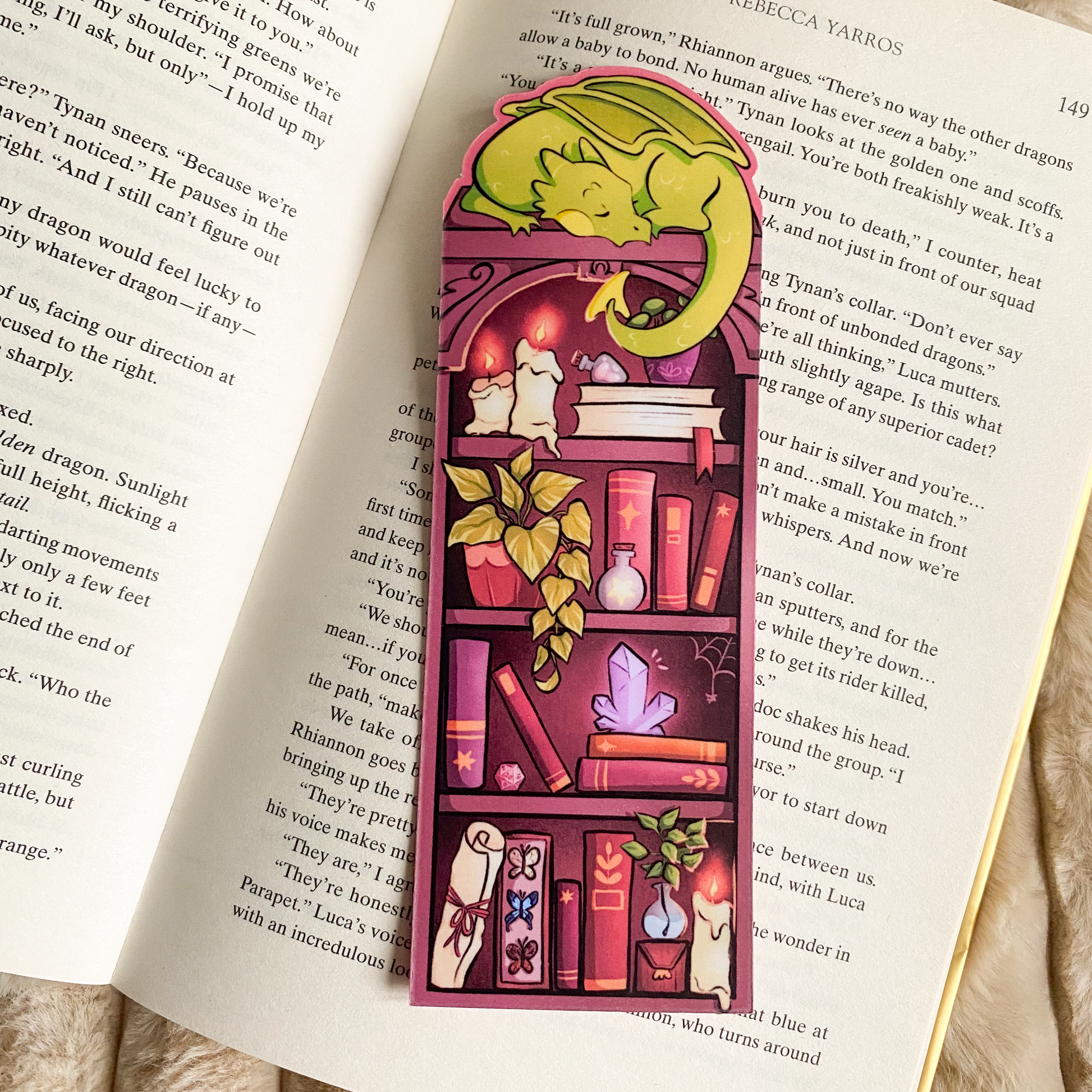 Picture of a fantasy dragon bookmark in a book. The dragon sleeps on top of the bookshelf that is filled with books and trinkets. 