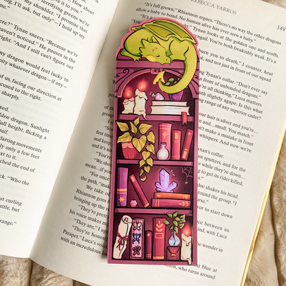 Picture of a fantasy dragon bookmark in a book. The dragon sleeps on top of the bookshelf that is filled with books and trinkets. 