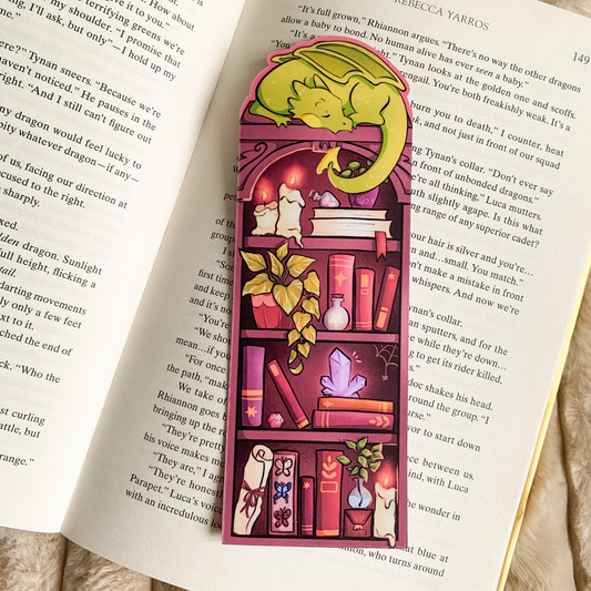 Picture of a fantasy dragon bookmark in a book. The dragon sleeps on top of the bookshelf that is filled with books and trinkets. 