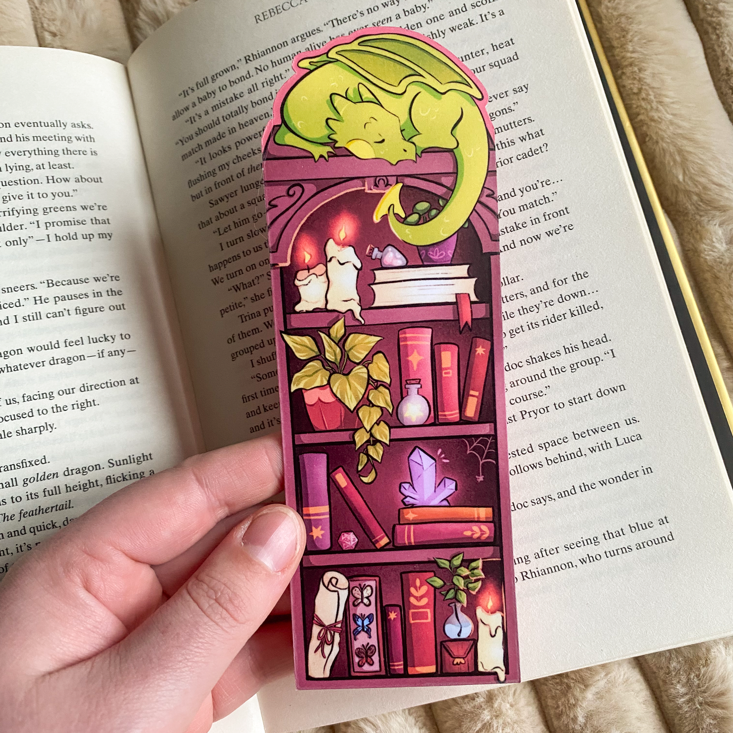 Picture of a fantasy dragon bookmark in a book. The dragon sleeps on top of the bookshelf that is filled with books and trinkets. 