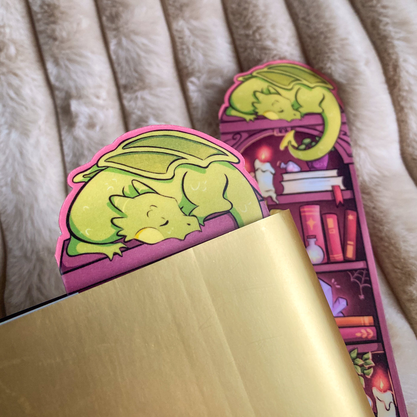 Picture of a fantasy dragon bookmark in a book. The dragon sleeps on top of the bookshelf that is filled with books and trinkets. 