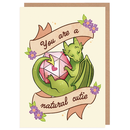 A product mockup of a D&D themed A6 postcard. It pictures a dragon with a D20 dice and text saying ''you are a natural cutie''