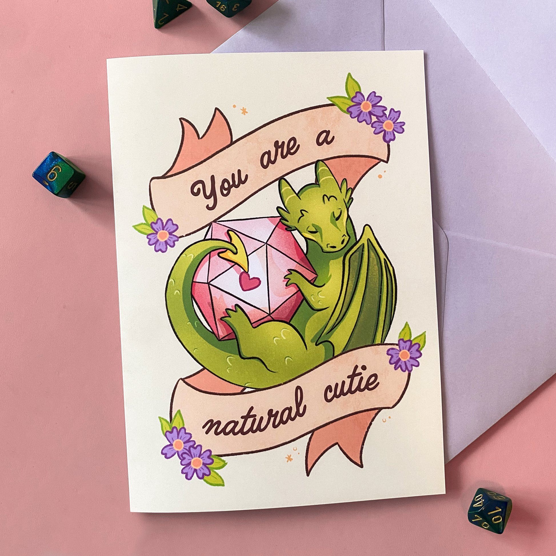 A product photo of a D&D themed postcard. It pictures a dragon with a D20 dice and text saying ''you are a natural cutie''
