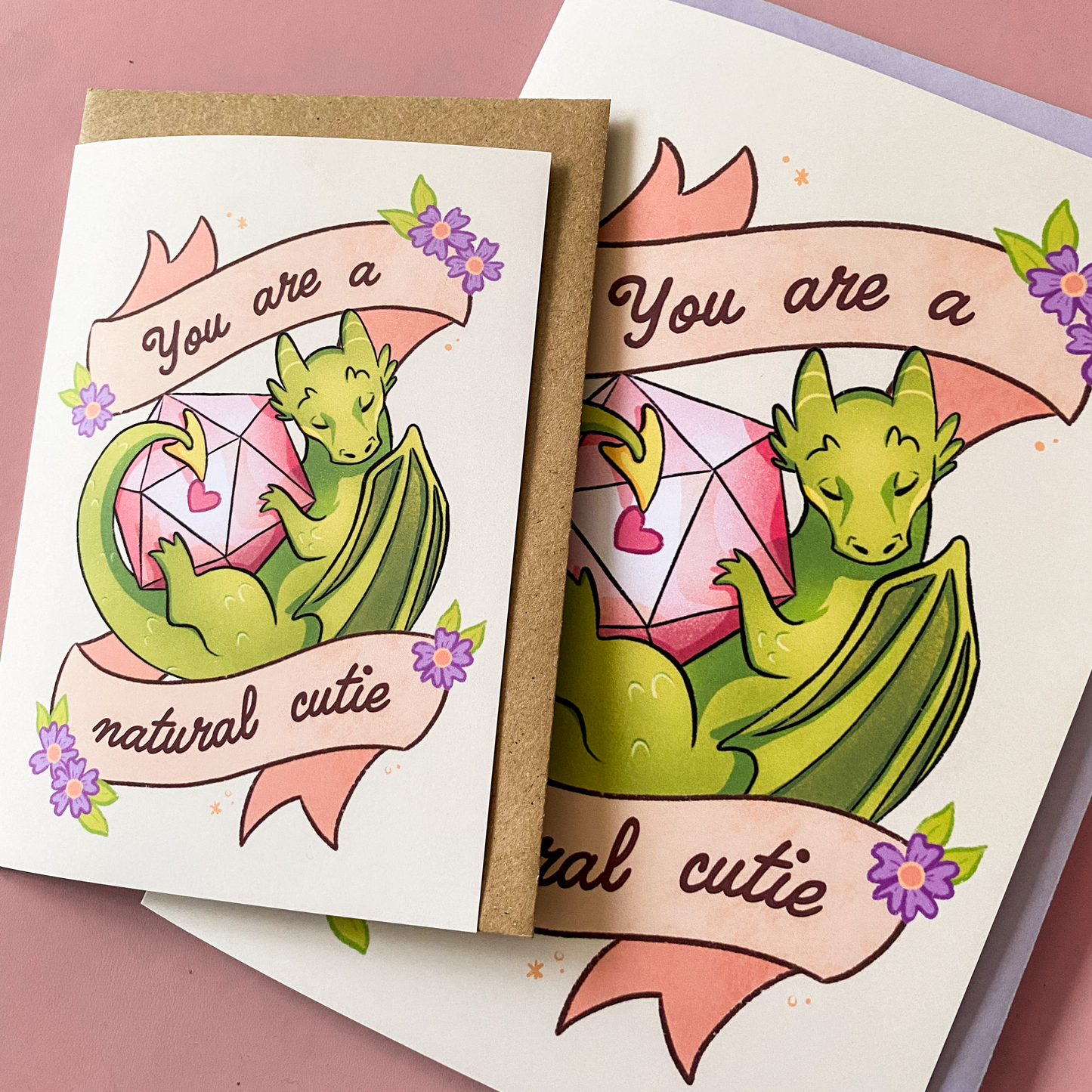 A product photo of a D&D themed postcard. It pictures a dragon with a D20 dice and text saying ''you are a natural cutie''