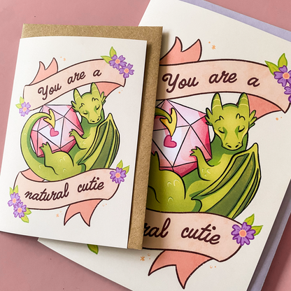 A product photo of a D&D themed postcard. It pictures a dragon with a D20 dice and text saying ''you are a natural cutie''