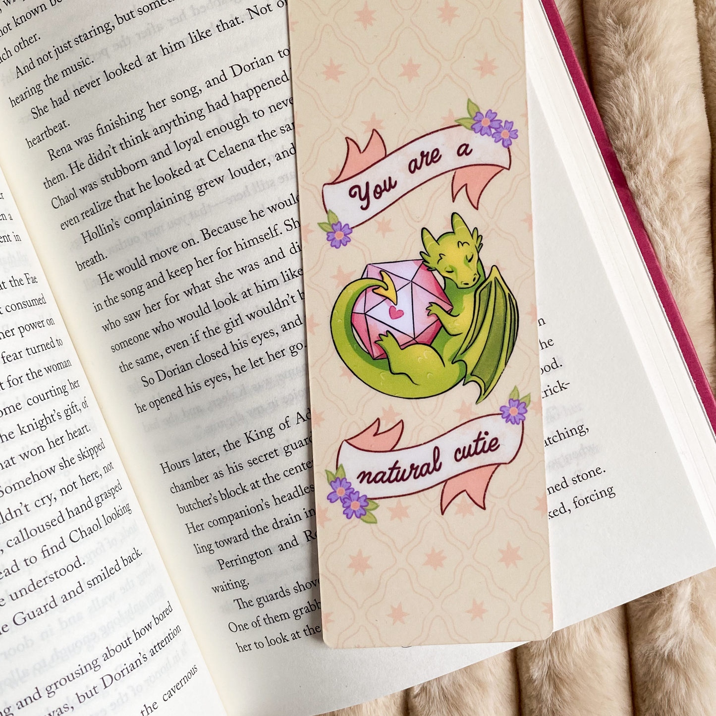 A product photo of a bookmark with an illustration of a green dragon holding a D20 dice. There is text saying ''you are a natural cutie''. The bookmark is laying in a book. 