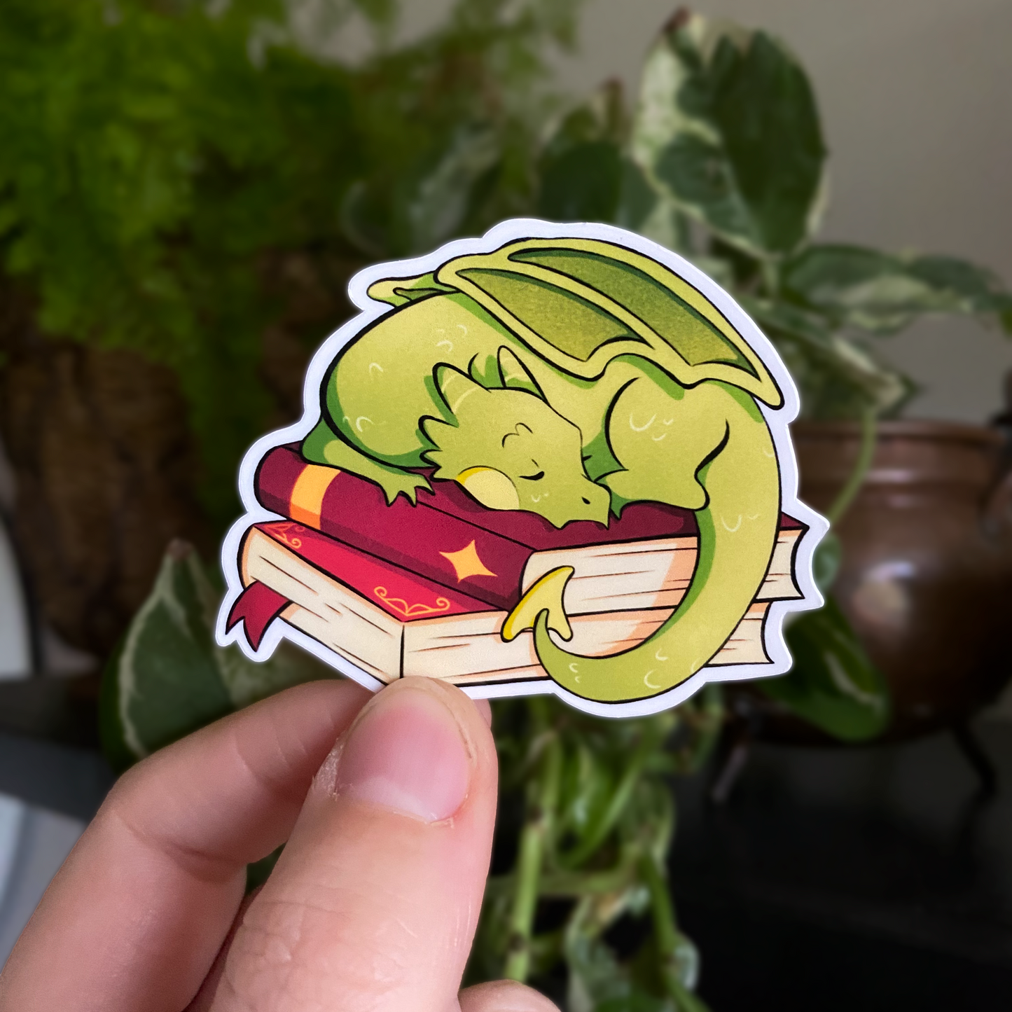 A productphoto of a fantasy themed vinyl sticker with a dragon sleeping on a stack of books. 