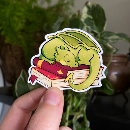 A productphoto of a fantasy themed vinyl sticker with a dragon sleeping on a stack of books. 