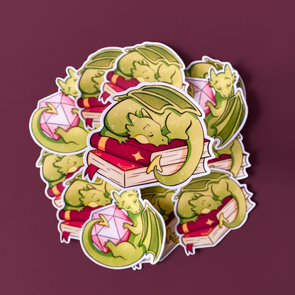 A productphoto of a fantasy themed vinyl sticker with a dragon sleeping on a stack of books. 